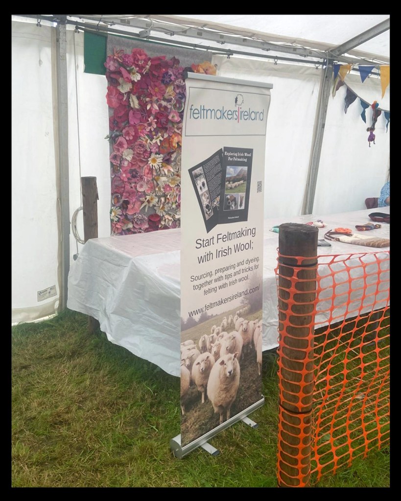 Poster of our book, 'Exploring Irish Wool for Felting' with sheep at the base. Next to that is a table with felted purses, and a backdrop of felted floral panel in pink.